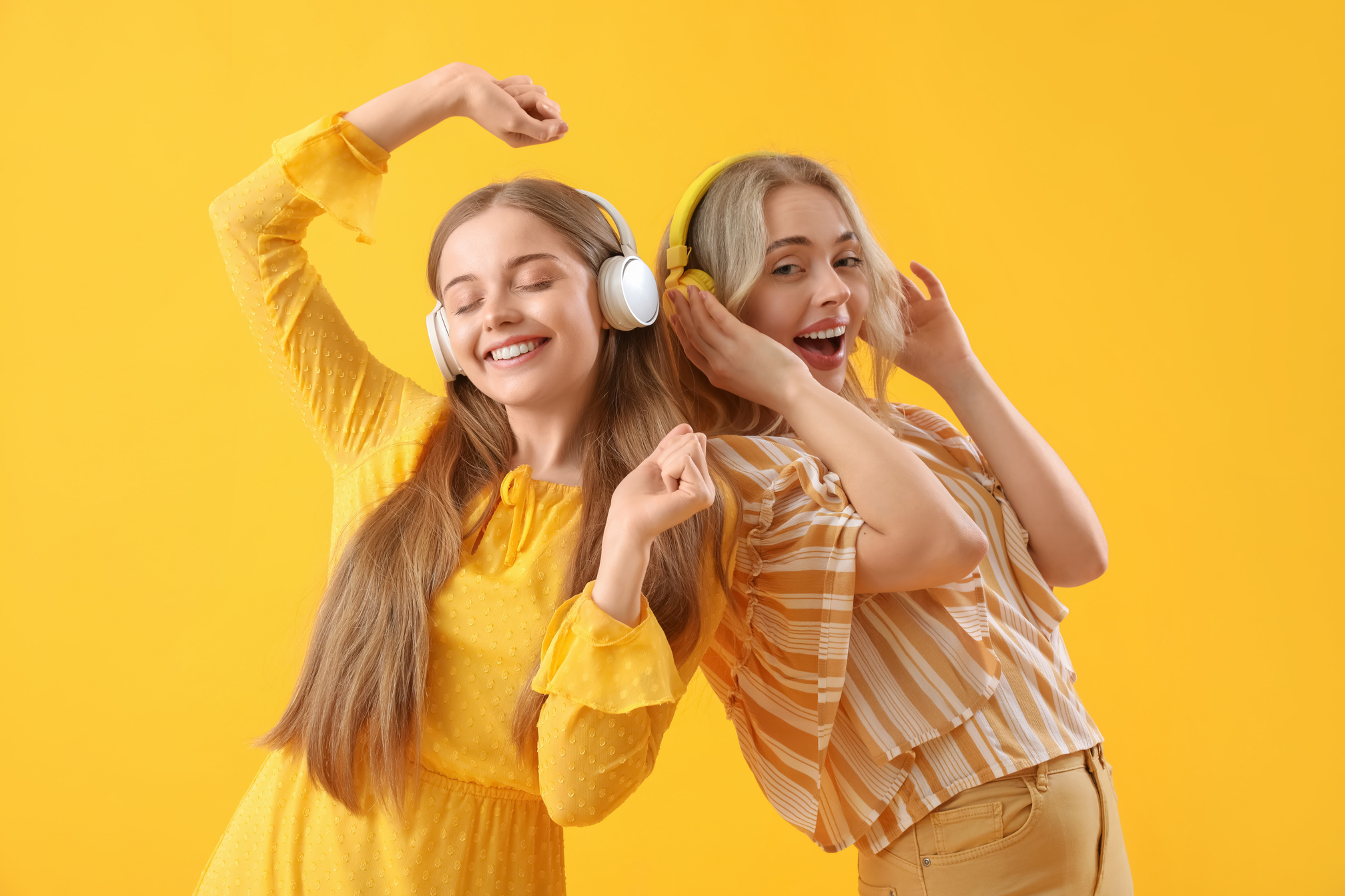 Young Sisters in Headphones Dancing on Yellow Background